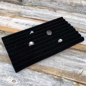 May include: Black velvet ring display with eight slots for rings. There are four rings on the display, one with a turquoise stone, one with a dark red stone, one with a pearl, and one with a clear stone.