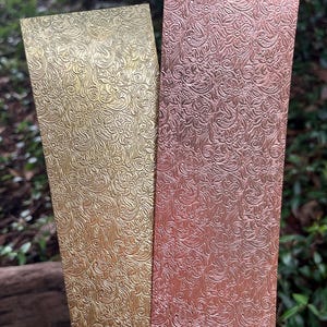 May include: Two rectangular metal panels with embossed floral patterns. One panel is gold, the other is a copper color. The panels are side by side, with a blurred green background.