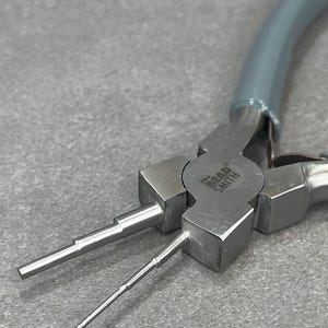 May include: Close-up of The Bead Smith round nose pliers. The pliers are silver-colored with gray handles. The tool has two round, tapered tips for jewelry making and wire wrapping.