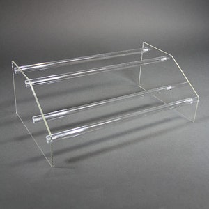 May include: A clear acrylic tiered display stand with three levels. The stand is designed to hold jewelry or other small items.