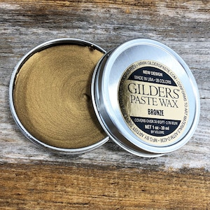 May include: Open tin of bronze-colored Gilders Paste Wax. The lid is next to the open tin, revealing the product's color and texture. The label reads "Bronze" and "Net 1 oz - 30 ml by volume." The product is designed for art materials.