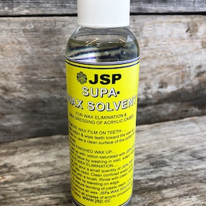 May include: A clear glass bottle with a yellow label that reads "JSP Supa. Wax Solvent". The label also includes instructions for using the solvent to remove wax from teeth and acrylic cases. The bottle is sitting on a wooden surface.