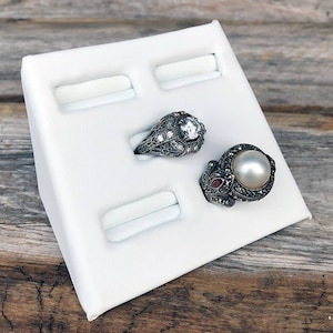 May include: Two ornate rings displayed on a white jewelry holder. One ring features a large, clear gemstone, while the other has a large pearl. The rings are set against a rustic wooden background.