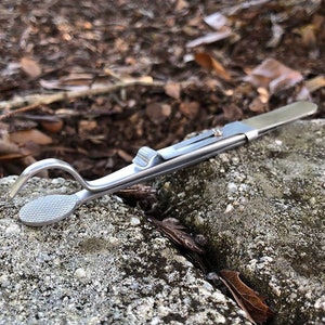 May include: A silver-toned tweezers with a curved tip and a textured grip. The tweezers are lying on a gray rock.