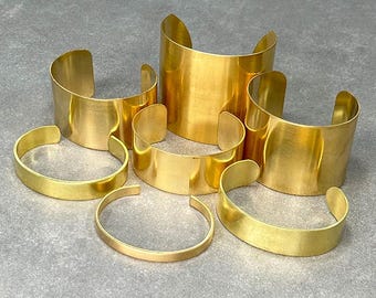 May include: A set of six gold-toned metal bracelets of varying widths. The bracelets are stacked on a gray surface.