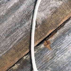 May include: A length of off-white, braided cotton rope, lying diagonally across weathered wooden planks. The rope has a slightly frayed end and a thicker, knotted end. The wood grain is visible, with variations in color from light to dark.