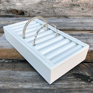 May include: White rectangular ring holder with eight slots for storing rings. Two silver rings are displayed in the holder.