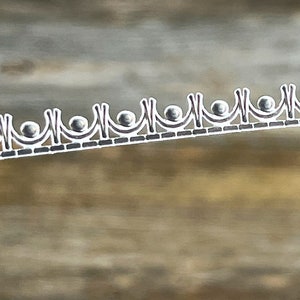 May include: A silver metal trim with a repeating pattern of small circles and arches. The trim is on a wood background.