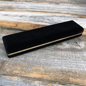 May include: A black velvet jewelry box with a gold trim. The box is closed and sitting on a wooden surface.