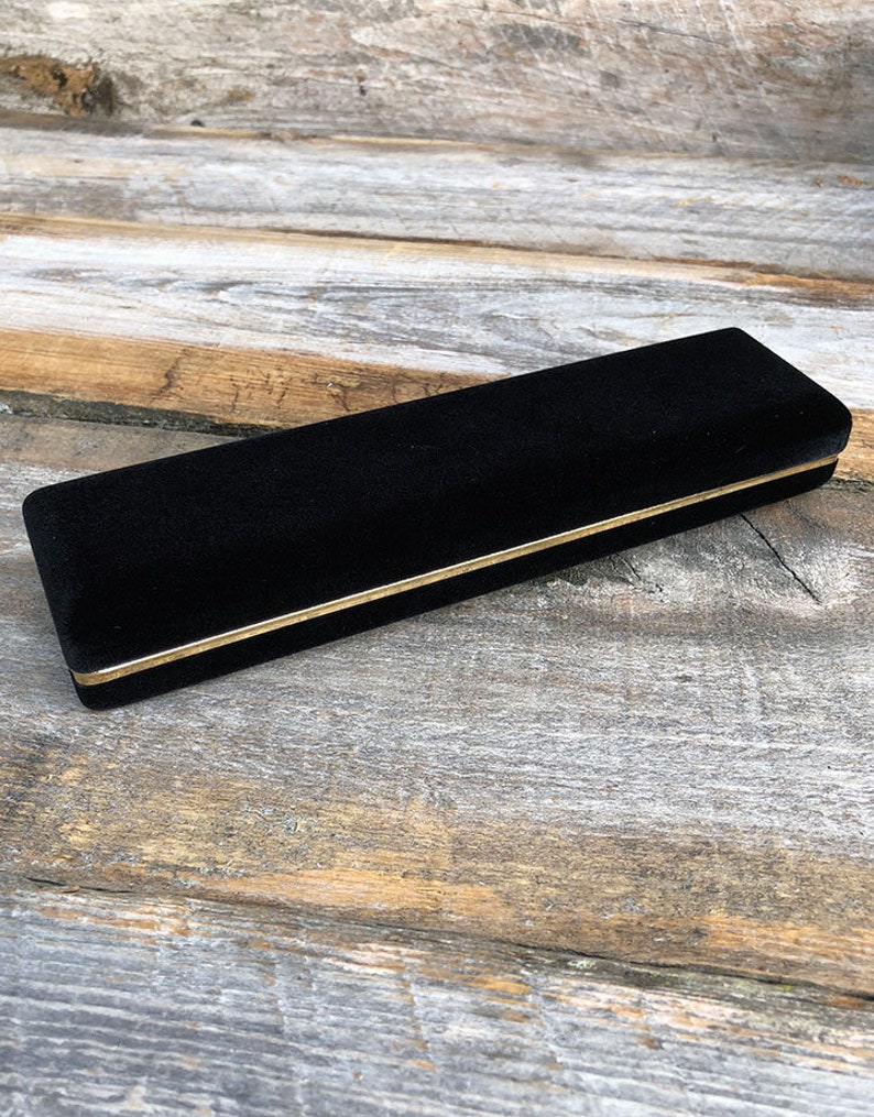 May include: A black velvet jewelry box with a gold trim. The box is closed and sitting on a wooden surface.