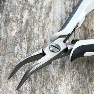 May include: Close-up of needle-nose pliers with curved jaws. The pliers have a silver metal finish with white and black handles. The tool is designed for precision work and is set against a weathered wooden surface.