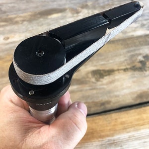May include: A black sanding tool with a gray sanding belt. The tool is held in a hand and has a round base with a small hole in the center.