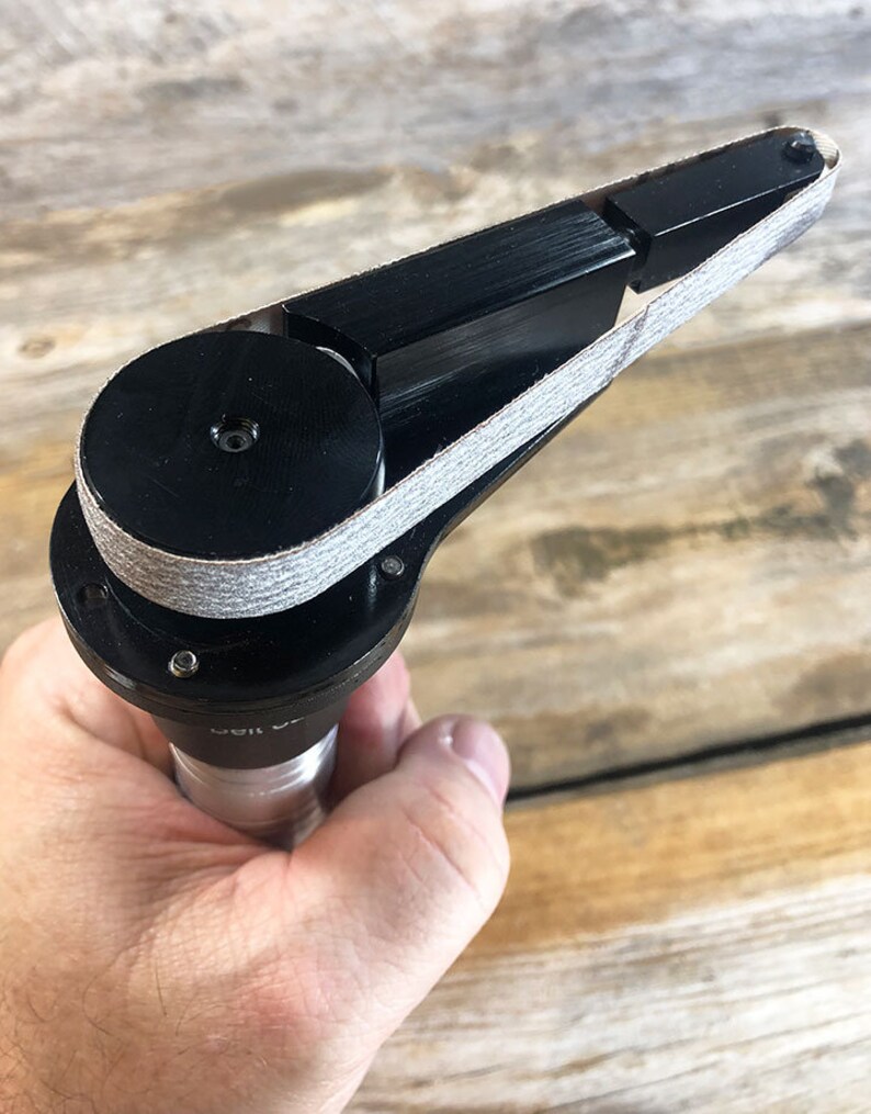 May include: A black sanding tool with a gray sanding belt. The tool is held in a hand and has a round base with a small hole in the center.