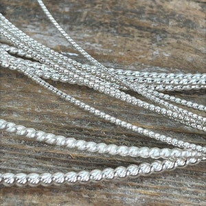 May include: A close-up of several silver chains of different styles and thicknesses. The chains are arranged in a layered pattern on a wooden surface.