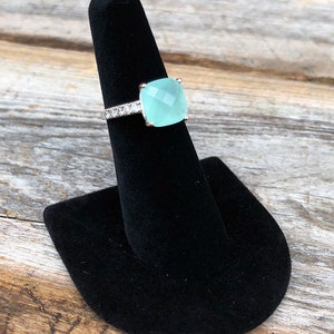 May include: A silver ring with a light blue square gemstone and a row of clear stones around the band.