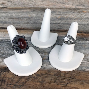 May include: Three white ring display stands with three rings on them. The rings are silver with different colored stones.