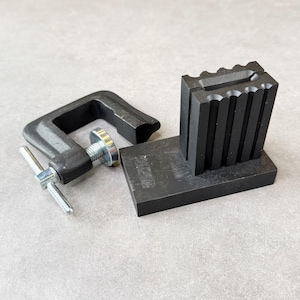 May include: A black metal C-clamp with a silver-colored threaded screw and T-handle, positioned next to a black rectangular metal block featuring a series of vertical, semi-circular grooves along one side and a central horizontal slot.