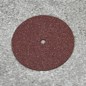 Puede incluir: Disco abrasivo circular de color granate con un agujero central. El disco tiene una superficie texturizada, probablemente para aplicaciones de lijado o esmerilado. El disco está sobre un fondo gris.