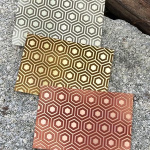 May include: Three rectangular metal sheets with a repeating geometric honeycomb pattern. The sheets are in shades of silver, gold, and copper. The sheets are displayed on a gray, textured surface.