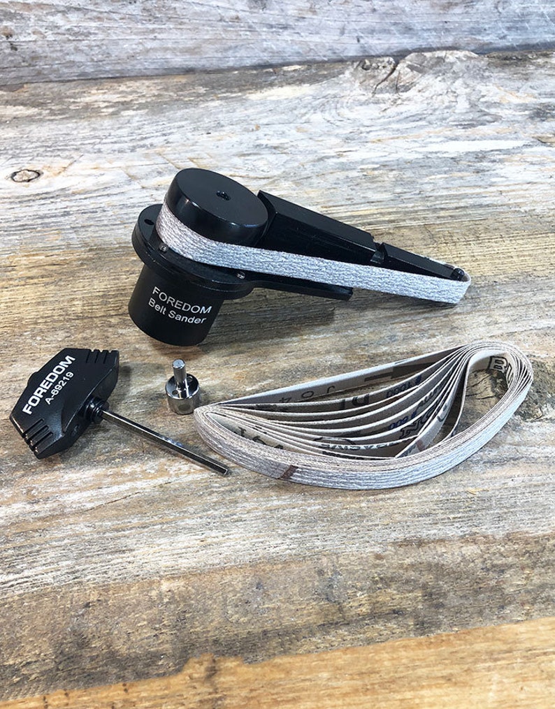 May include: A black Foredom A88219 belt sander with a gray sanding belt. The sander is on a wooden surface with a set of sanding belts next to it. There is also a black wrench and a metal rod.