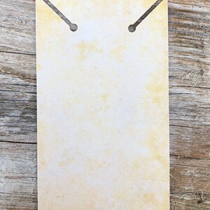 Pode incluir: Um cart&atilde;o de exibi&ccedil;&atilde;o retangular de papel com uma textura manchada em amarelo p&aacute;lido e branco, semelhante a pergaminho. A borda superior possui duas fendas angulares que levam a orif&iacute;cios circulares para prender pe&ccedil;as de joalharia.