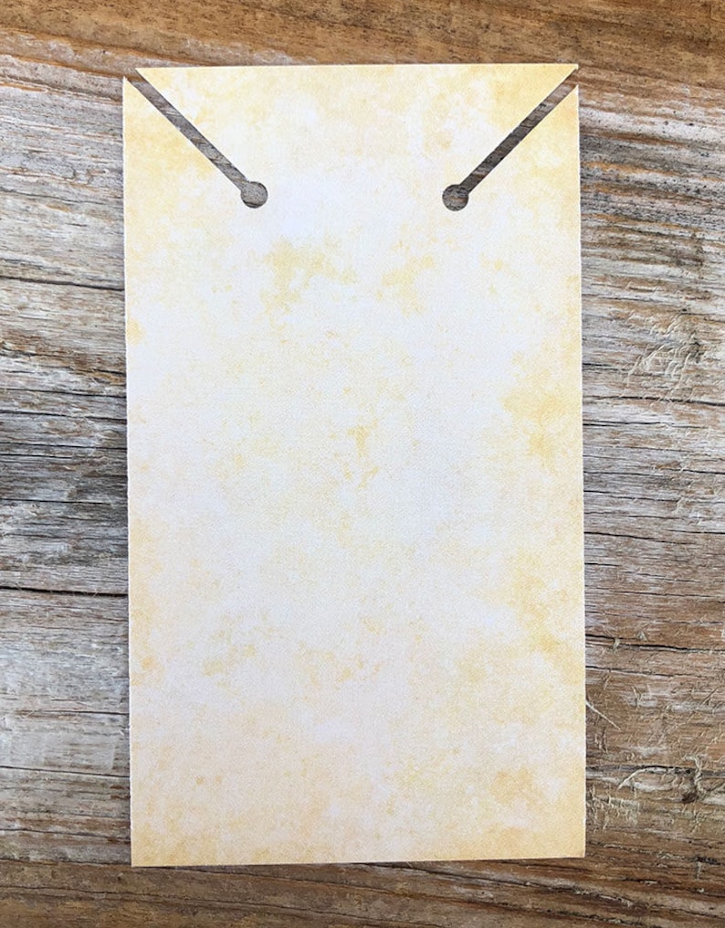 Pode incluir: Um cart&atilde;o de exibi&ccedil;&atilde;o retangular de papel com uma textura manchada em amarelo p&aacute;lido e branco, semelhante a pergaminho. A borda superior possui duas fendas angulares que levam a orif&iacute;cios circulares para prender pe&ccedil;as de joalharia.
