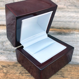 May include: A dark mahogany wood ring box with a white velvet interior. The box is open and the lid is propped up.