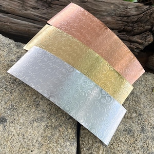 May include: Four metal bands in silver, gold, copper, and bronze, each with a repeating swirl pattern. The bands are arranged diagonally on a stone surface, showcasing their different colors and textures. The bands are likely for jewelry making.