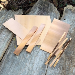 May include: Various sizes of copper sheets and strips are arranged on a weathered wooden surface. The copper pieces have a warm, reddish-brown hue and reflect light, showcasing their smooth, polished surfaces. The arrangement suggests materials for crafting or metalworking projects.