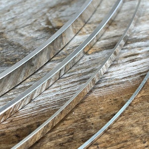 May include: Four silver metal strips, each with a slightly rounded edge, are stacked on top of each other on a wooden surface. The strips are all the same width, but they vary in length.