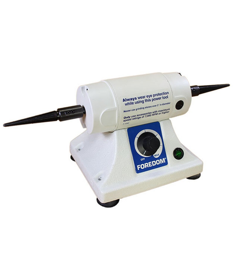 Może przedstawiać: Białe narzędzie elektryczne Foredom z czarną wrzecioną i pokrętłem regulacji prędkości. Narzędzie ma zieloną lampkę kontrolną i etykietę ostrzegawczą z napisem "Zawsze noś ochronę oczu podczas korzystania z tego narzędzia elektrycznego. Nigdy nie używaj kamieni szlifierskich o średnicy większej niż 2 cale. Używaj tylko akcesori&oacute;w o maksymalnej prędkości 7 000 obr./min lub wyższej."