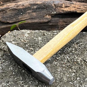May include: A close-up of a hammer with a wooden handle. The hammer has a silver head and a rounded striking face. The hammer is lying on a gray, textured surface.