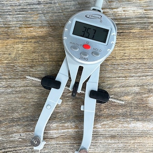 May include: A white digital caliper with black accents and a display reading 3.57 mm. The caliper has a zero button and a hold button. The caliper is on a wooden surface.
