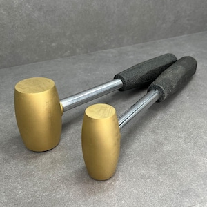 May include: Two gold-colored metal hammers with black rubber handles. The hammers are lying on a gray surface.
