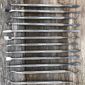 May include: An assortment of stainless steel dental tools, including probes, spatulas, and excavators, arranged on a weathered wooden surface. The tools have textured handles for grip and various working ends for different dental procedures.