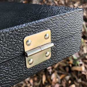 K&ouml;nnte beinhalten: Nahaufnahme einer schwarzen Box mit einem silbernen Scharnier. Die Box hat eine strukturierte Oberfl&auml;che.