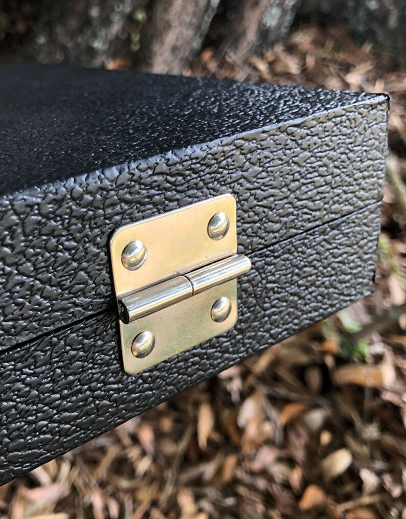 K&ouml;nnte beinhalten: Nahaufnahme einer schwarzen Box mit einem silbernen Scharnier. Die Box hat eine strukturierte Oberfl&auml;che.