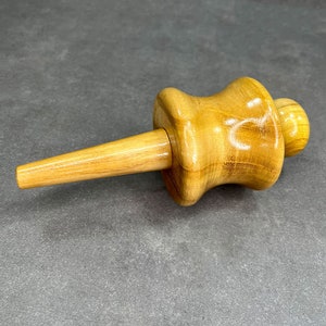 May include: A wooden bottle stopper with a light brown finish. The stopper has a cylindrical stem, a bulbous midsection, and a small, rounded top. The wood grain is visible, and the stopper appears to be handcrafted.