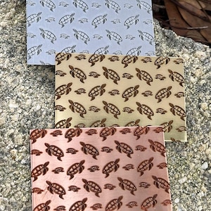 May include: Three rectangular metal plates with a repeating pattern of turtles. The plates are in silver, gold, and copper colors. The turtles are etched into the metal, creating a textured surface. The plates are displayed on a rough, stone surface.