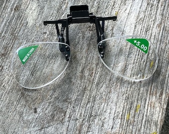 以下が含まれることがあります： クリップオン式の老眼鏡、+5.00の拡大鏡付き。透明なレンズには「MADE IN CHINA」と書かれた緑色のステッカーが付いています。黒いフレームには、既存のメガネに取り付けるためのクリップが付いています。メガネは風化した木製の表面に置かれています。