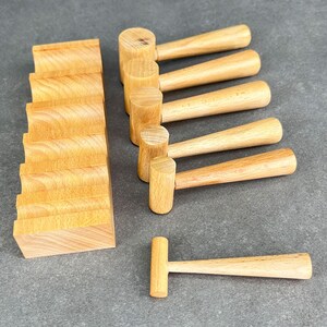 May include: A collection of small, light-colored wooden mallets and a wooden block with a curved design. The mallets have rounded heads and tapered handles. The items are arranged on a gray surface.