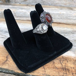 May include: Two silver rings with gemstones on a black velvet ring display stand. The ring on the left has a large round clear stone and the ring on the right has a large round red stone.
