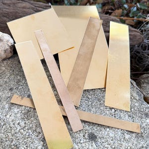 May include: A collection of brass metal pieces in various shapes, including rectangles and strips. The pieces have a shiny, golden appearance, with some showing signs of oxidation. The arrangement is on a textured, gray stone surface, with greenery in the background.
