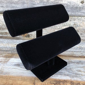 May include: Black velvet jewelry display stand with two tiers for bracelets or watches. The stand has a square base and a cylindrical center post.