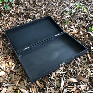 K&ouml;nnte beinhalten: Eine schwarze rechteckige Box mit einer strukturierten Oberfl&auml;che und einem Scharnierdeckel. Die Box ist ge&ouml;ffnet und das Innere ist mit schwarzem Stoff ausgekleidet.