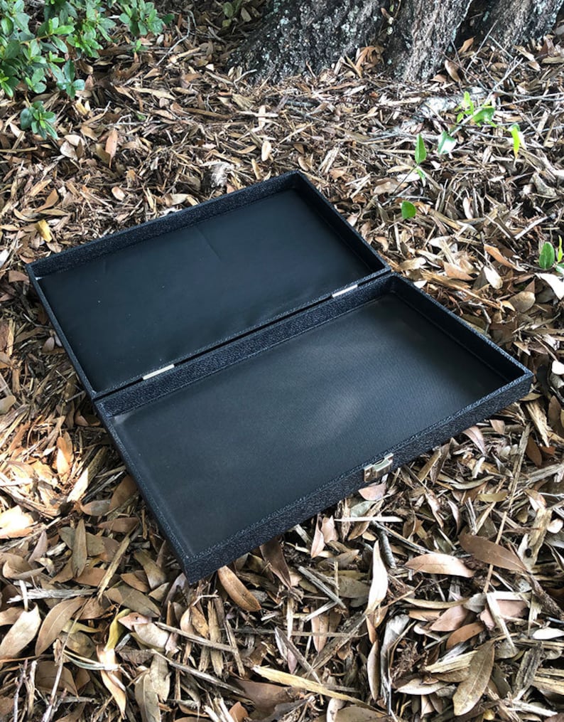 K&ouml;nnte beinhalten: Eine schwarze rechteckige Box mit einer strukturierten Oberfl&auml;che und einem Scharnierdeckel. Die Box ist ge&ouml;ffnet und das Innere ist mit schwarzem Stoff ausgekleidet.
