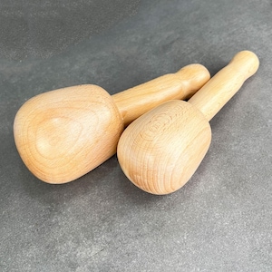 May include: Two wooden meat mallets with rounded heads and long handles. The mallets are made of light-colored wood and are positioned on a gray surface. The mallets are designed for tenderizing meat.
