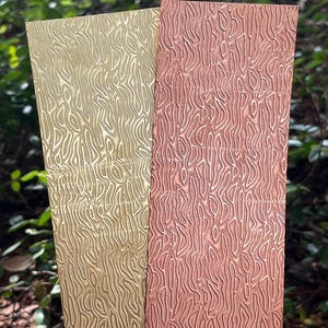 May include: Two rectangular metal sheets with a textured, organic pattern. One is a warm gold color, and the other is a reddish-brown. The sheets are side-by-side, with the textured surfaces facing the viewer. The background is blurred green foliage.
