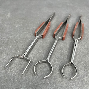May include: Three stainless steel tweezers with wooden handles. The tweezers have different tip styles: a forked tip, a rounded tip, and a pointed tip.