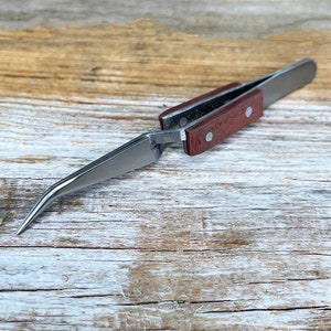 May include: A pair of stainless steel tweezers with a curved tip and a wooden handle. The tweezers are lying on a wooden surface.